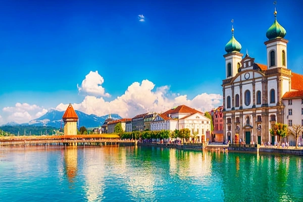 Iconic Chapel Bridge and Jesuit Church on the banks of Lake Lucerne with the Swiss Alps in the background.