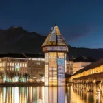 Chapel Bridge (Kapellbrücke) and Water Tower illuminated at night in Lucerne, Switzerland with the Swiss Alps in the background, reflecting on Lake Lucerne.