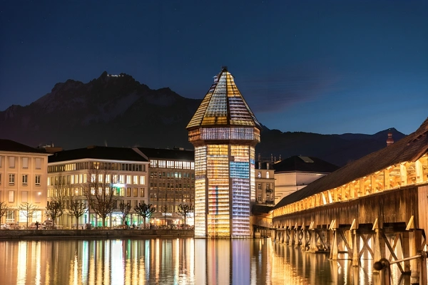 Chapel Bridge (Kapellbrücke) and Water Tower illuminated at night in Lucerne, Switzerland with the Swiss Alps in the background, reflecting on Lake Lucerne.