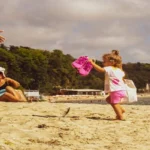 Happy toddler playing on a sandy beach in Cornwall while family relaxes nearby, representing a family-friendly beach holiday in the UK