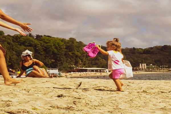 Happy toddler playing on a sandy beach in Cornwall while family relaxes nearby, representing a family-friendly beach holiday in the UK
