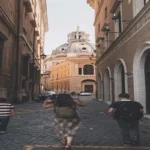 Visitors walking through a narrow cobblestone street in Rome with a historic church dome in the background