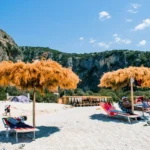 Gjipe Beach Albania featuring straw umbrellas, white sand, and towering canyon cliffs under a blue sky.