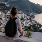 Solo traveler sitting on a viewpoint overlooking a coastal hillside town at sunset with mountains and sea in background